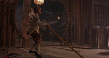 Movie still from “Iron Monkey” (1993), directed by Woo-Ping Yuen – A man in a white shirt and brown pants holding a wooden stick; Wide shot, Low angle