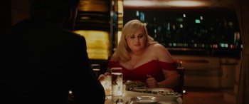 Movie still from “Isn't It Romantic” (2019), directed by Todd Strauss-Schulson – A woman sitting at a table with a plate of food; Medium shot, Over the shoulder angle