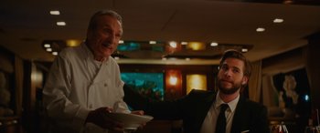 Movie still from “Isn't It Romantic” (2019), directed by Todd Strauss-Schulson – A man in a suit is handing a plate to a man in a chef's uniform; Medium shot, Low angle