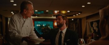 Movie still from “Isn't It Romantic” (2019), directed by Todd Strauss-Schulson – A man wearing a suit and tie is talking to another man in a suit; Medium shot, Over the shoulder angle