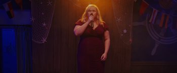 Movie still from “Isn't It Romantic” (2019), directed by Todd Strauss-Schulson – A woman in a red dress is singing into a microphone; Wide shot, Over the shoulder angle