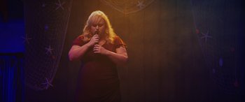 Movie still from “Isn't It Romantic” (2019), directed by Todd Strauss-Schulson – A woman in a red dress is holding a microphone; Medium shot, Over the shoulder angle