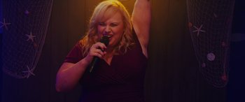 Movie still from “Isn't It Romantic” (2019), directed by Todd Strauss-Schulson – A woman holding a microphone up to her head; Medium shot, Low angle