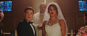 Movie still from “Isn't It Romantic” (2019), directed by Todd Strauss-Schulson – A man and a woman standing next to each other in front of a priest; Medium shot, Over the shoulder angle