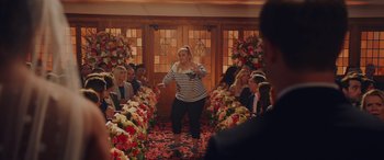 Movie still from “Isn't It Romantic” (2019), directed by Todd Strauss-Schulson – A woman standing in front of a group of people in a room; Wide shot, Over the shoulder angle