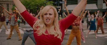 Movie still from “Isn't It Romantic” (2019), directed by Todd Strauss-Schulson – A woman in a red shirt is standing in front of a group of people; Medium shot, Over the shoulder angle