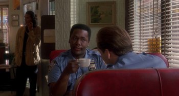 Movie still from “It Could Happen to You” (1994), directed by Andrew Bergman – A police officer sitting in front of another officer holding a coffee cup; Medium shot, Over the shoulder angle