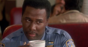 Movie still from “It Could Happen to You” (1994), directed by Andrew Bergman – A police officer holding a cup of coffee while sitting in a booth; Close Up shot, Over the shoulder angle