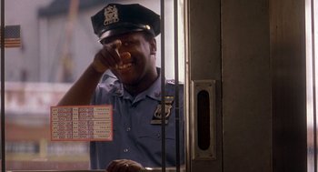 Movie still from “It Could Happen to You” (1994), directed by Andrew Bergman – A police officer eating a donut while standing in front of a window; Medium shot, Low angle