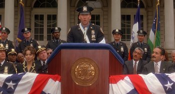 Movie still from “It Could Happen to You” (1994), directed by Andrew Bergman – A man in a police uniform standing at a podium; Medium shot, Low angle