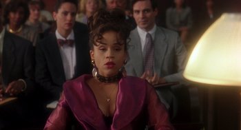 Movie still from “It Could Happen to You” (1994), directed by Andrew Bergman – A woman in a purple dress sitting in front of a group of people; Medium shot, Over the shoulder angle