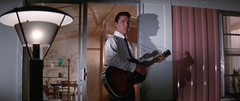 Movie still from “It Happened at the World's Fair” (1963), directed by Norman Taurog – A man holding a guitar while standing next to a door; Medium shot, Low angle