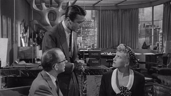 Movie still from “It Should Happen to You” (1954), directed by George Cukor – A group of people in a room with a building in the background; Medium shot, Over the shoulder angle