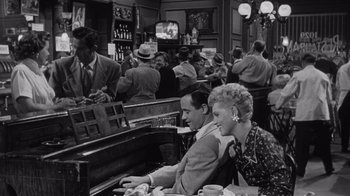 Movie still from “It Should Happen to You” (1954), directed by George Cukor – A man and a woman sitting at a table in front of a piano; Wide shot, High angle
