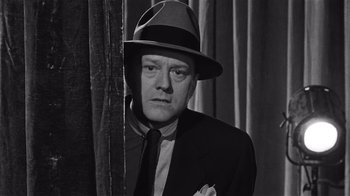 Movie still from “It Should Happen to You” (1954), directed by George Cukor – A man wearing a suit and a hat is leaning against a wall; Close Up shot, High angle