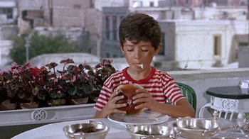 Movie still from “It Started in Naples” (1960), directed by Melville Shavelson – A young boy sitting at a table eating a hamburger; Medium shot, High angle