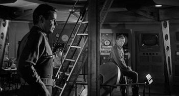 Movie still from “It! The Terror from Beyond Space” (1958), directed by Edward L. Cahn – A man sitting on top of a chair next to another man; Medium shot, Over the shoulder angle
