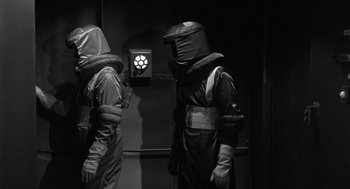 Movie still from “It! The Terror from Beyond Space” (1958), directed by Edward L. Cahn – Two people in black suits and helmets are standing in front of a wall; Medium shot, Low angle