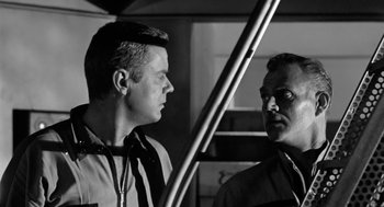 Movie still from “It! The Terror from Beyond Space” (1958), directed by Edward L. Cahn – Two men standing next to each other in front of a metal pole; Close Up shot, Low angle