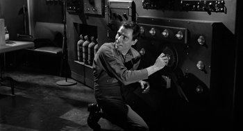 Movie still from “It! The Terror from Beyond Space” (1958), directed by Edward L. Cahn – A black and white photo of a man working on a machine; Medium shot, Low angle