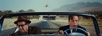 Movie still from “It's a Mad Mad Mad Mad World” (1963), directed by Stanley Kramer – Two people in a car driving down a road with a helicopter in the background; Medium shot, Over the shoulder angle