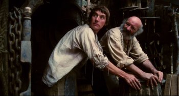 Movie still from “Jabberwocky” (1977), directed by Terry Gilliam – A man leaning against a wall next to another man with a beard; Medium shot, Low angle