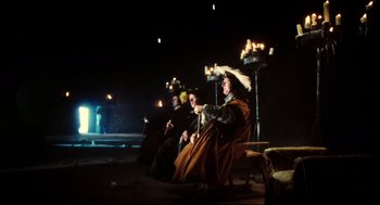 Movie still from “Jabberwocky” (1977), directed by Terry Gilliam – A group of people sitting on a stage with lit candles; Wide shot, Low angle