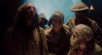 Movie still from “Jabberwocky” (1977), directed by Terry Gilliam – A group of people standing around each other in a room; Medium shot, Low angle