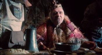 Movie still from “Jabberwocky” (1977), directed by Terry Gilliam – An old man sitting at a table in front of a pot of food; Medium shot, Low angle