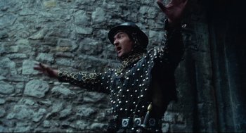 Movie still from “Jabberwocky” (1977), directed by Terry Gilliam – A man in a black and gold outfit standing next to a stone wall; Medium shot, Low angle