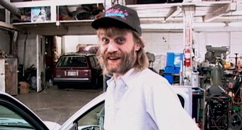 Movie still from “Jackass: The Movie” (2002), directed by Jeff Tremaine – A man in a baseball cap standing in front of a car; Medium shot, High angle