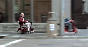 Movie still from “Jackass: The Movie” (2002), directed by Jeff Tremaine – An older man riding a scooter down a street; Wide shot, High angle
