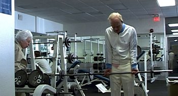 Movie still from “Jackass: The Movie” (2002), directed by Jeff Tremaine – An older man in a white shirt is lifting a barbell in a gym; Wide shot, Low angle
