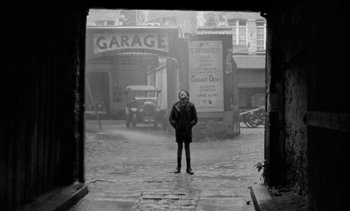 Movie still from “Jacquot of Nantes” (1991), directed by Agnès Varda – A man standing on a street in front of a garage; Wide shot, High angle