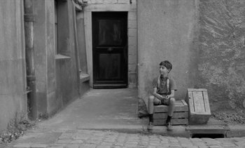 Movie still from “Jacquot of Nantes” (1991), directed by Agnès Varda – A young boy sitting on top of a wooden crate; Wide shot, High angle