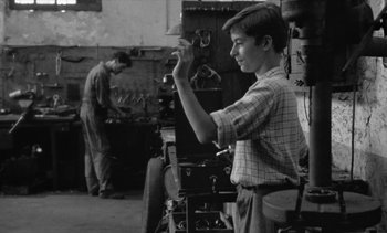 Movie still from “Jacquot of Nantes” (1991), directed by Agnès Varda – A young man in plaid shirt holding up a tool; Medium shot, High angle
