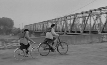Movie still from “Jacquot of Nantes” (1991), directed by Agnès Varda – Two people riding bicycles on a road near a bridge; Wide shot, High angle