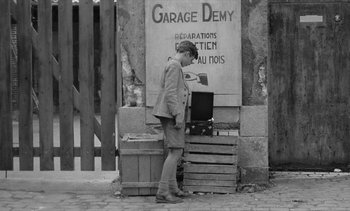 Movie still from “Jacquot of Nantes” (1991), directed by Agnès Varda – A man standing in front of crates on the sidewalk; Wide shot, High angle