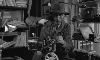 Movie still from “Jacquot of Nantes” (1991), directed by Agnès Varda – A man in a hat is working on an old movie projector; Medium shot, High angle