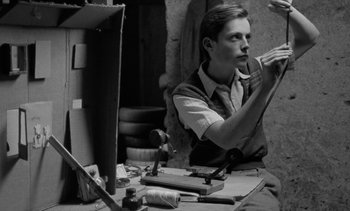 Movie still from “Jacquot of Nantes” (1991), directed by Agnès Varda – A man sitting at a table with a pair of scissors; Medium shot, Low angle