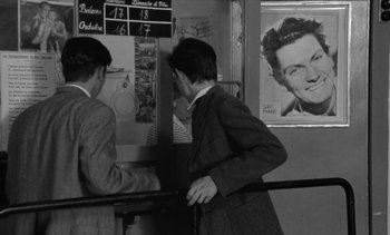 Movie still from “Jacquot of Nantes” (1991), directed by Agnès Varda – Two young men are standing in front of a poster of a man's face; Medium shot, Over the shoulder angle