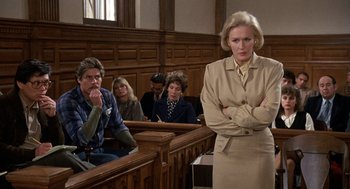 Movie still from “Jagged Edge” (1985), directed by Richard Marquand – A woman standing in front of a group of people in a court room; Medium shot, Over the shoulder angle