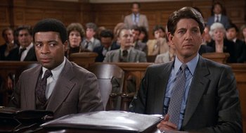 Movie still from “Jagged Edge” (1985), directed by Richard Marquand – Two men in suits sit in front of a crowd of onlookers in a courtroom; Medium shot, Low angle