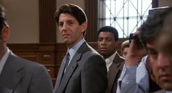 Movie still from “Jagged Edge” (1985), directed by Richard Marquand – A man in a suit and tie standing in front of other people; Close Up shot, Over the shoulder angle