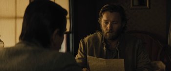 Movie still from “Jane Got a Gun” (2015), directed by Gavin O'Connor – A man sitting at a table looking at a piece of paper; Medium shot, Over the shoulder angle