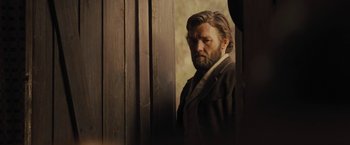 Movie still from “Jane Got a Gun” (2015), directed by Gavin O'Connor – A man with long hair and a beard standing next to a wooden wall; Medium shot, Over the shoulder angle