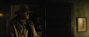 Movie still from “Jane Got a Gun” (2015), directed by Gavin O'Connor – A man wearing a hat standing in a dark room; Medium shot, Over the shoulder angle