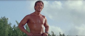 Movie still from “Jaws 3-D” (1983), directed by Joe Alves – A man with no shirt is standing in the grass; Medium shot, Low angle