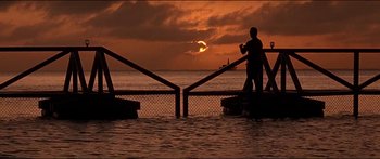 Movie still from “Jaws 3-D” (1983), directed by Joe Alves – A man standing on a pier near the ocean at sunset; Extreme Wide shot, Low angle