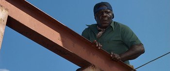 Movie still from “Jaws: The Revenge” (1987), directed by Joseph Sargent – A man in a green shirt and a blue hat is holding a metal beam; Close Up shot, Low angle